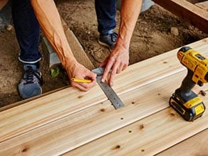 Two hands mark a wooden beam with a pencil against a metal ruler for a cut, with a drill resting on the beam nearby.