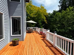 Wooden deck attached to a gray house with white railing, potted plants, and outdoor seating under an umbrella, overlooking trees.
