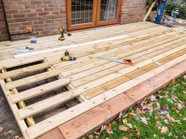 deck-builder-san-marcos-tx-Texas-1 | Ambassador Construction Construction of a wooden deck underway: exposed joists, planks laid out, and tools (drill, saw, level) on the surface near a brick house.