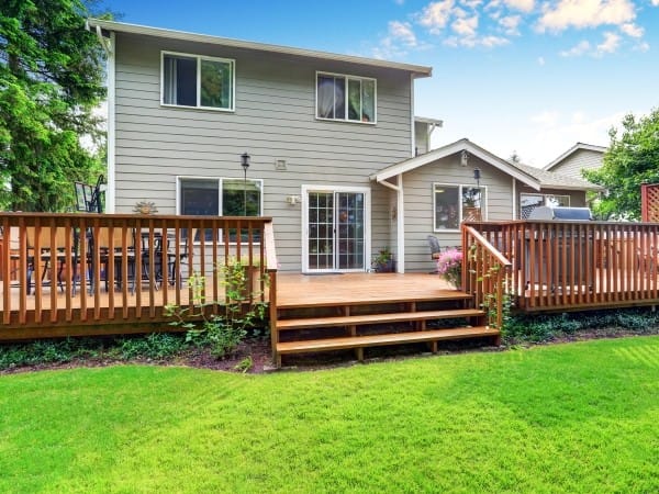 deck-builder-san-marcos-tx-Texas-2 | Ambassador Construction Two-story gray house with a large wooden deck and stairs, glass doors opening to the deck, and a green lawn behind.