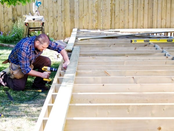 Man kneeling in a plaid shirt drills a wooden deck frame in a backyard, with a level and tools nearby.